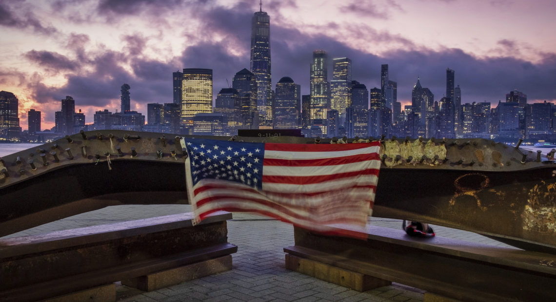 A U.S. Flag hanging from a steal girder, damaged in the Sept. 11, 2001 attacks on the World Trade Center, blows in the breeze at a memorial in Jersey City, N.J., Sept. 11, 2019 as the sun rises behind the One World Trade Center building and the re-developed area where the Twin Towers of World Trade Center once stood in New York City on the 18th anniversary of the attacks.