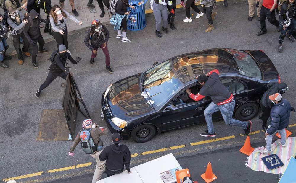 A man drives into the crowd at 11th and Pike, injuring at least one person, before exiting the car and brandishing an apparent firearm, Sunday, June 7, 2020, in Seattle, during protests over the death of George Floyd in Minneapolis. The man reaching into the car to stop it was injured. CREDIT: Rutz/The Seattle Times via AP