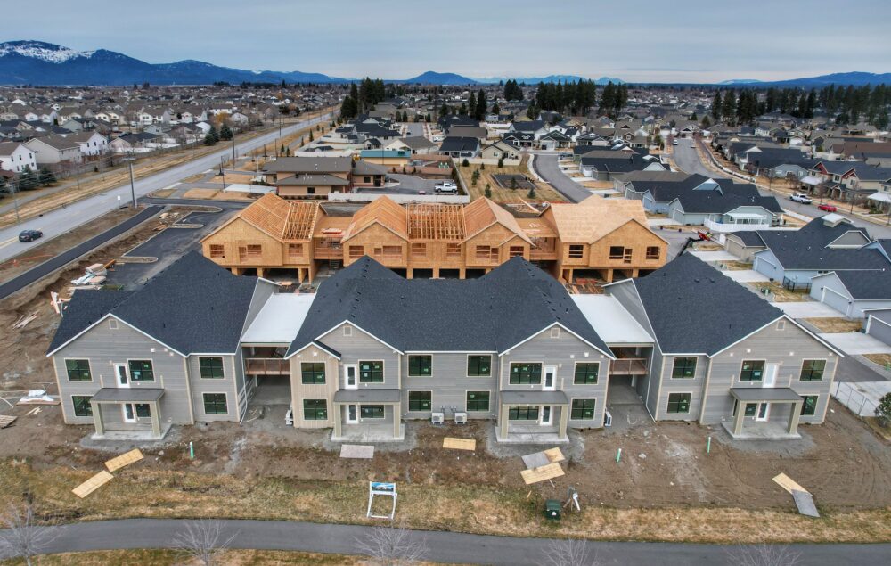 A newly constructed multi-home complex in shades of grey stands before a wooden house frame still under construction.