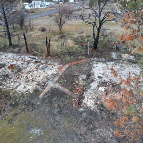 Charred ruins in grey ash sit on wet ground surrounded by trees.