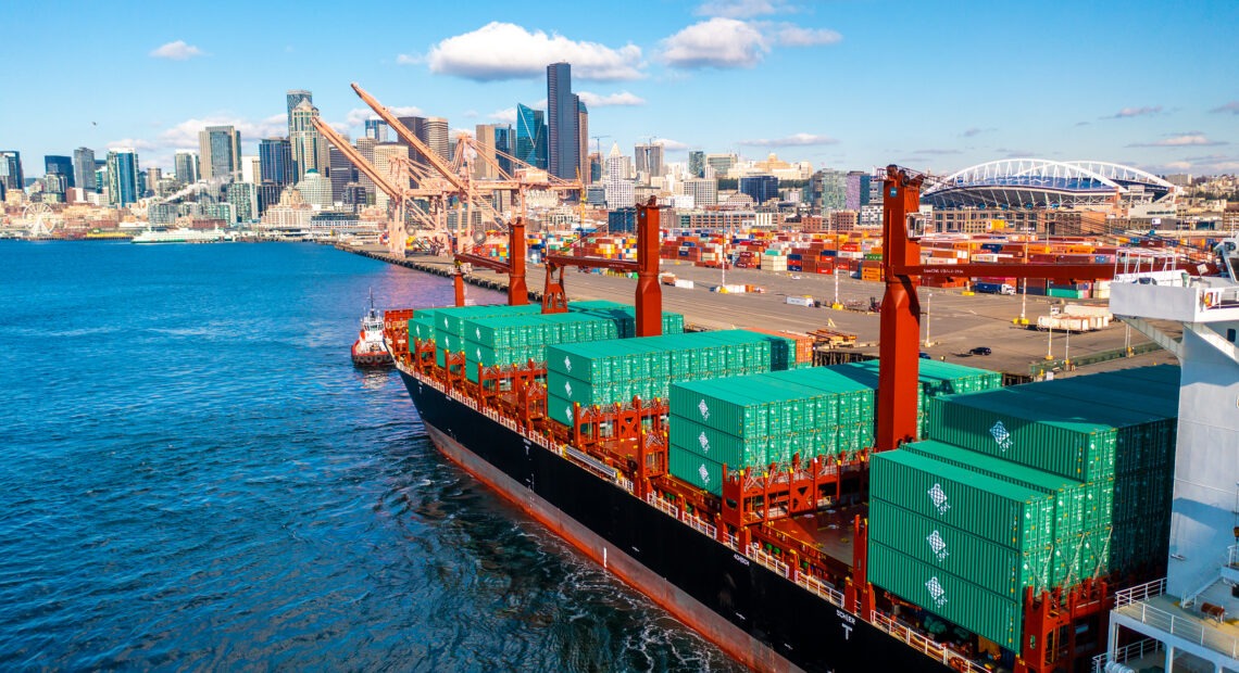 A bright red cargo ship with teal green containers makes its way across the blue ocean to the port of Seattle.