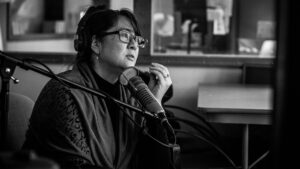 Sueann Ramella sits slightly forward in her chair in the studio, talking to Jiemei Lin, who isn't pictured and sits somewhere to the right of the shot. Sueann's hand is close to her face, gesturing, and she wears an empathetic look.