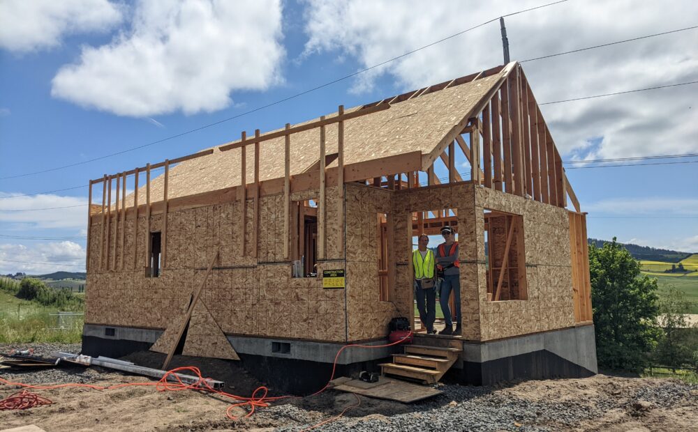 Two women in construction helmets stand in the frame of an unfinished house against a blue sky surrounded by green fields.