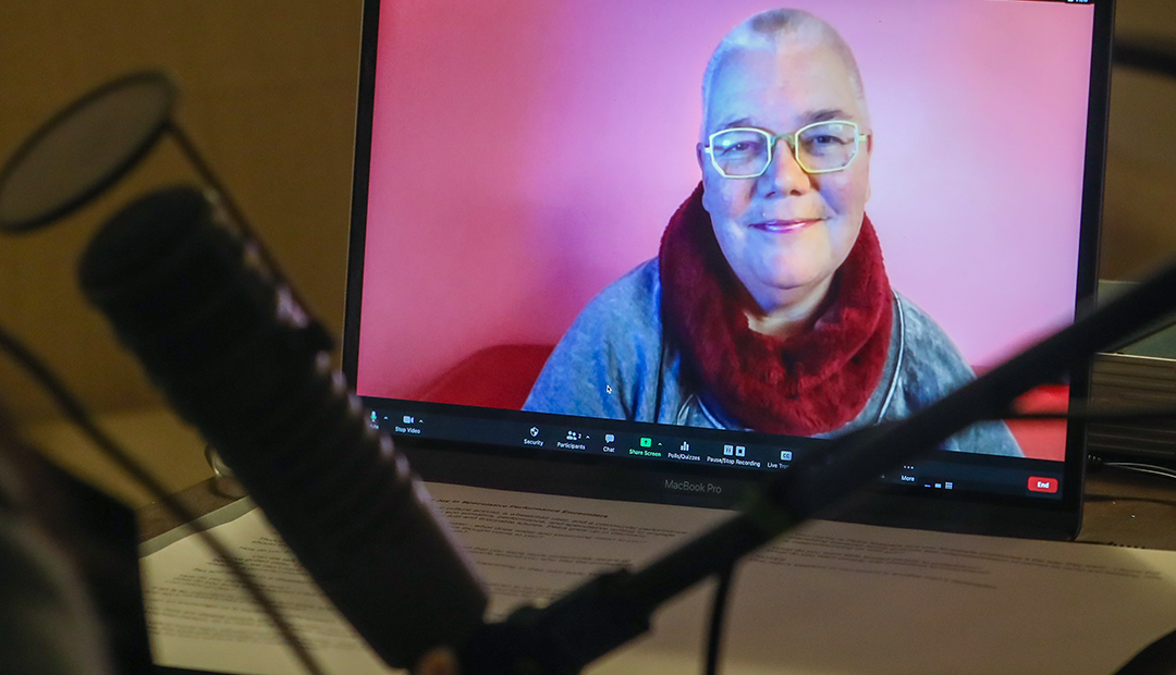 Petra Kuppers is seen through a screen. She is in front of a pink background. Sueann sits at the computer. A microphone is in view.