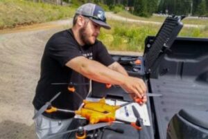 A man in a black t-shirt and blue baseball cap holds onto an orange drone.