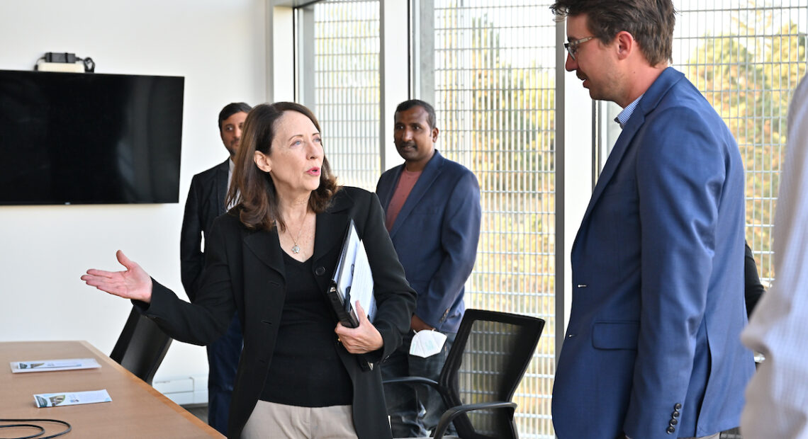 Senator Maria Cantwell in beige pants and a black suit stands in a conference room and talks to a man in a blue jacket.