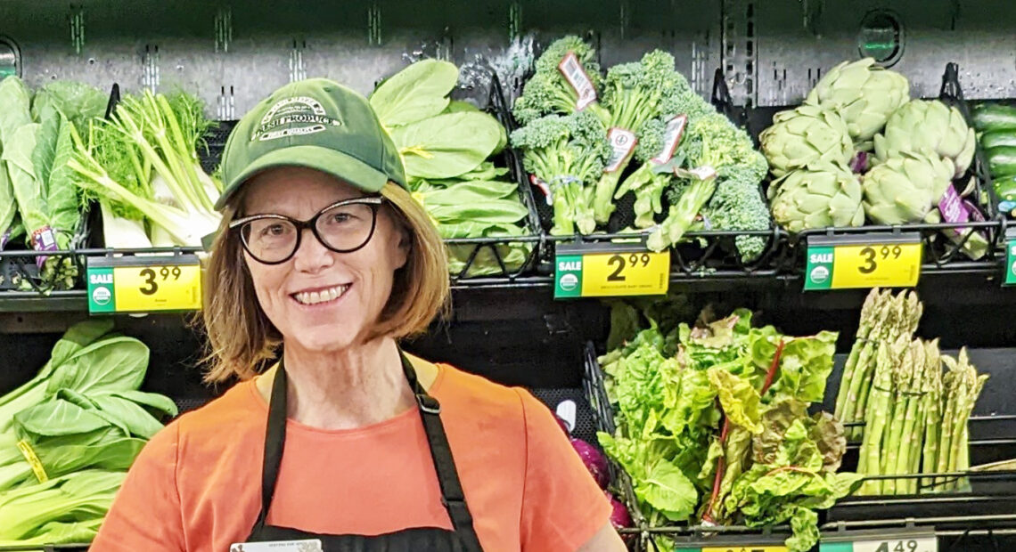 A women with red hair, black glasses, and orange shirt and a green hat poses in front of a produce section at Fred Meyer in Richland, Washington.