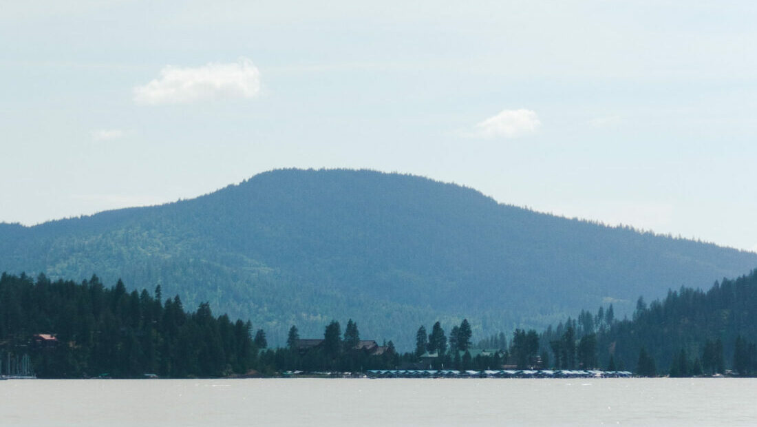 A blue mountain rises beyond a dark green treeline along a blue lake.