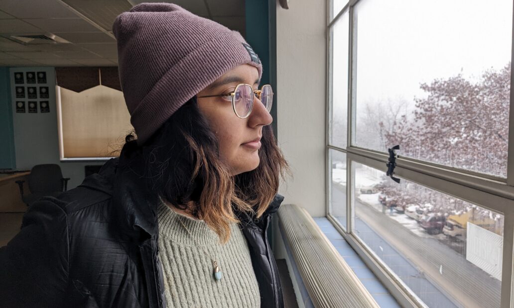 A woman with tan skin, glasses, and a purple beanie stares out the window into a snowy Moscow, Idaho.