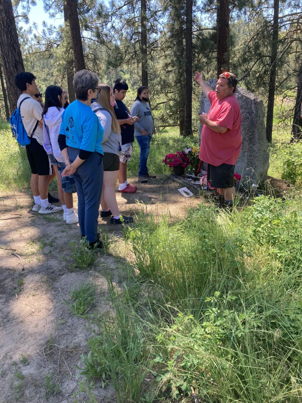 A man in a red shirt speaks to a crowd of students under green trees. 