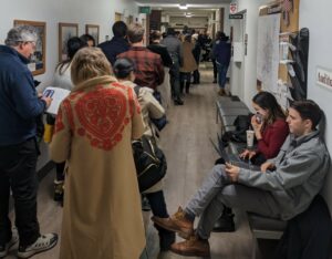 A white hallway with wood floors is filled with reporters lining up to enter a courtroom. Two are sitting on a brown bench, and a woman in a red coat is on the phone.