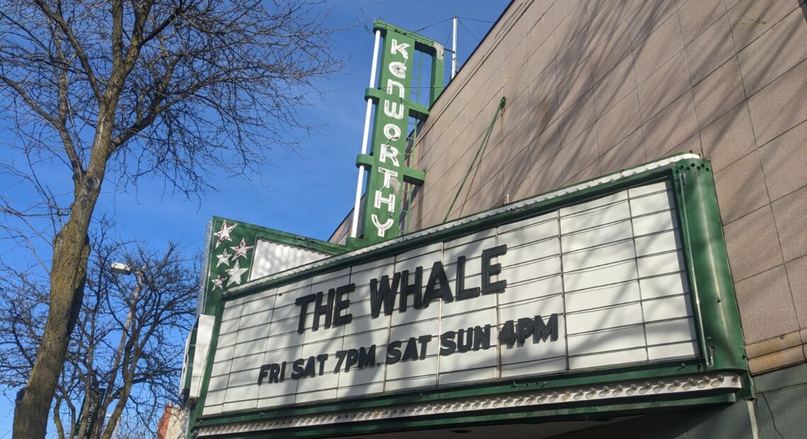 A white marquis spells out THE WHALE in black lettering on the front of an old theater with a green sign that reads "Kenworthy" against a blue sky.