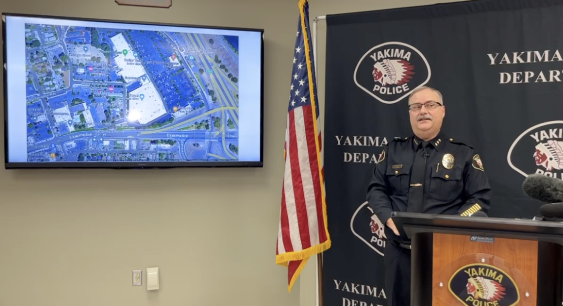 A police officer in uniform wearing glasses stands behind a podium and in front of a background reading "Yakima Police" on his left is an American flag and a computer screen showing a map of where the suspect was found dead.