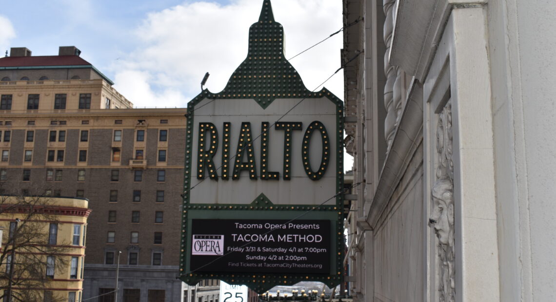 A photo of the marquee outside the Tacoma Opera's premiere of the Tacoma Method opera shows a green rimmed marquee with the word "Rialto" in large block letters. There are office buildings in the background and the signs hangs above a street. In the far background is a blue sky with bright, white clouds.