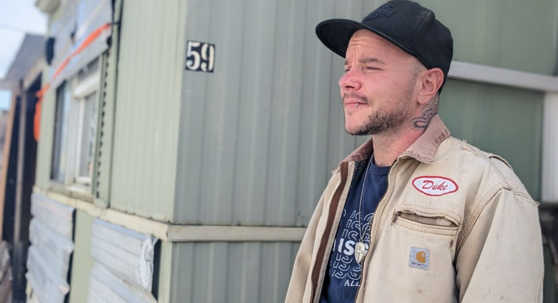 A man with a neck tattoo and a beige jacket stands in front of a green manufactured home in Latah County, Idaho.