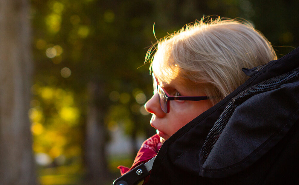 A woman with short, blond hair sits facing toward the left in profile of the camera and looking through dark-rimmed glasses. In the background are green trees out of focus in golden sunlight.