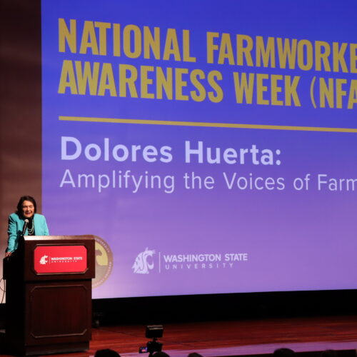 Dolores Huerta speaks at a podium at Washington State University. She wears a black shirt and a turquoise jacket.