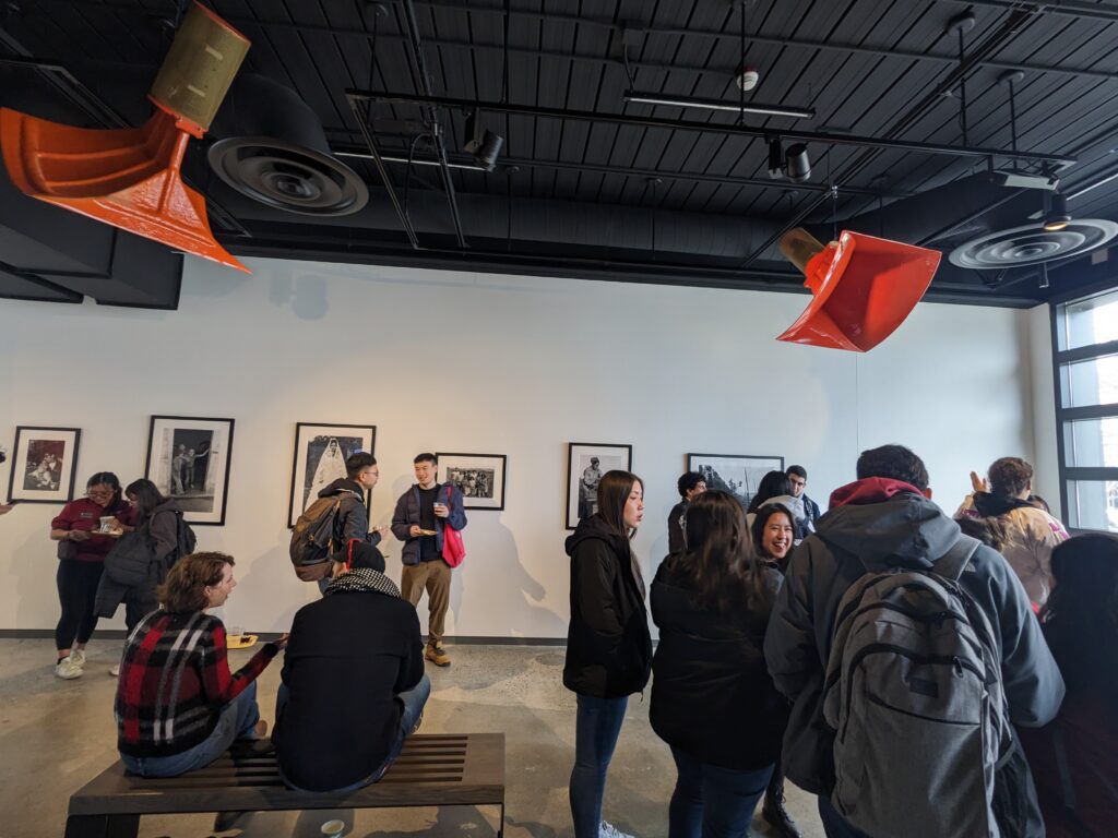 Students in coats and backpacks socialize in a room of white walls with black and white framed photographs on display.