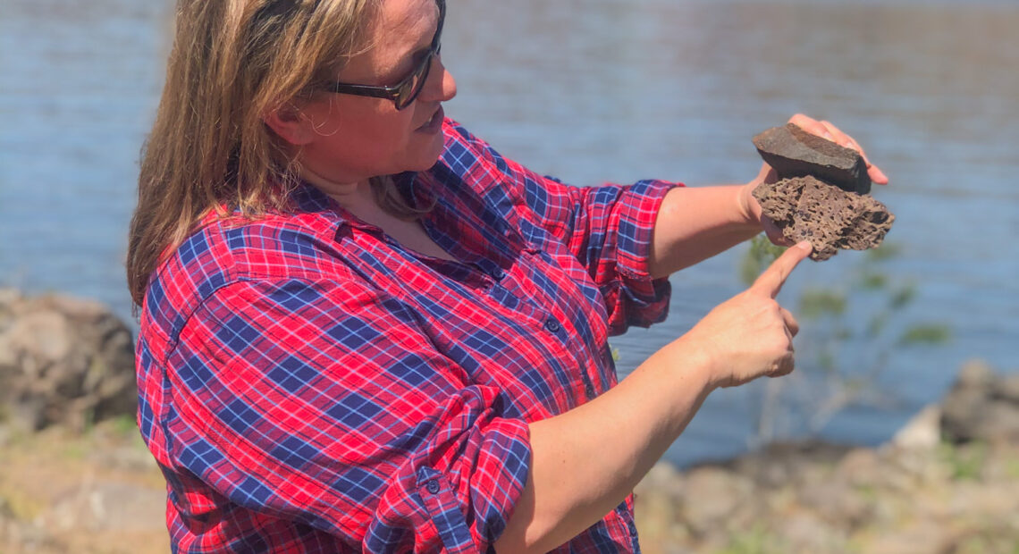 A women in a blue and red plaid shirt stands in front of a blue river. She is holding two gray rocks on top of each other. in the background, on the other side of the river, there are rocky hills and a blue sky.