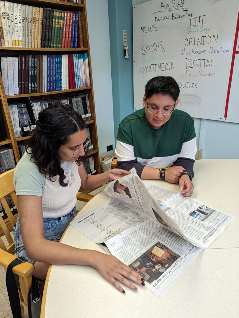 Two young people sit at a desk in the Argonaut office reading an article from the campus paper. 