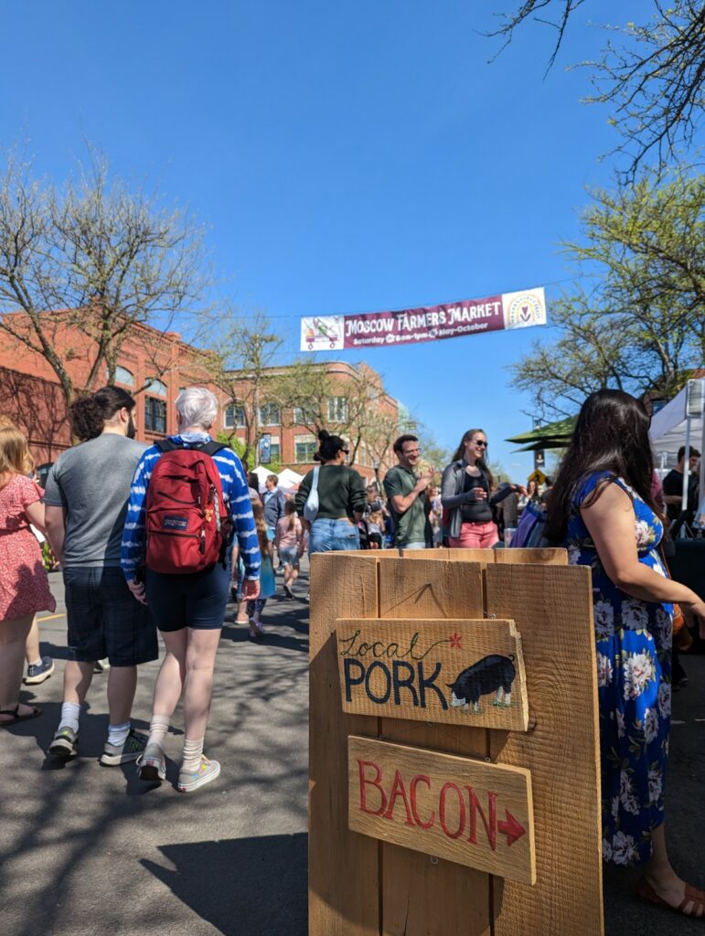 A banner reading "Moscow Farmer's Market" is hung between trees under a blue sky as Main Street is filled with people.