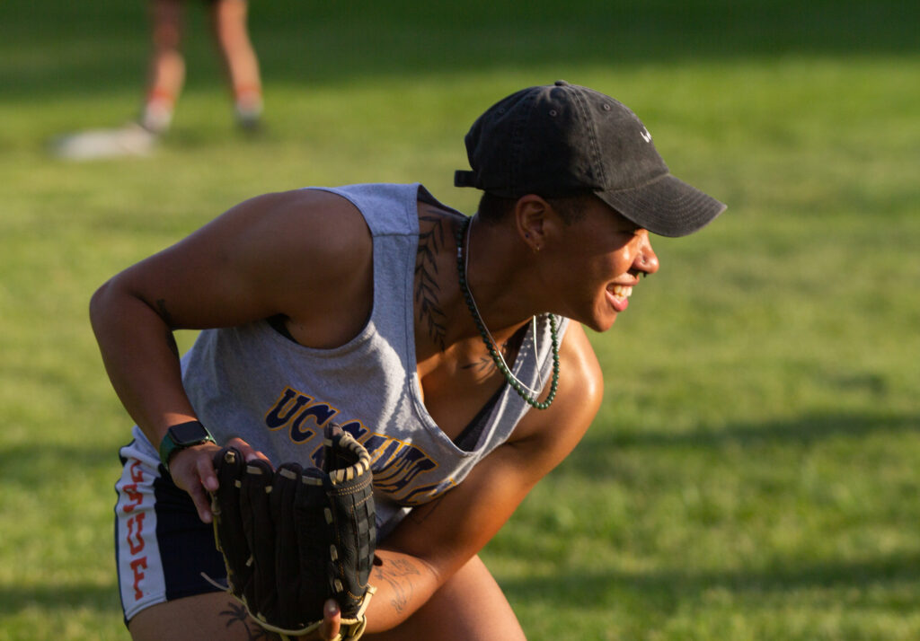 A young baseball player wearing a black ball cap grins toward the right of frame. They wear two necklaces, one silver metal and one green bead, and a botanical tattoo of leaves encircle their neck. They crouch, with a baseball mitt in one hand. In the background a field of blurred out grass is visible.