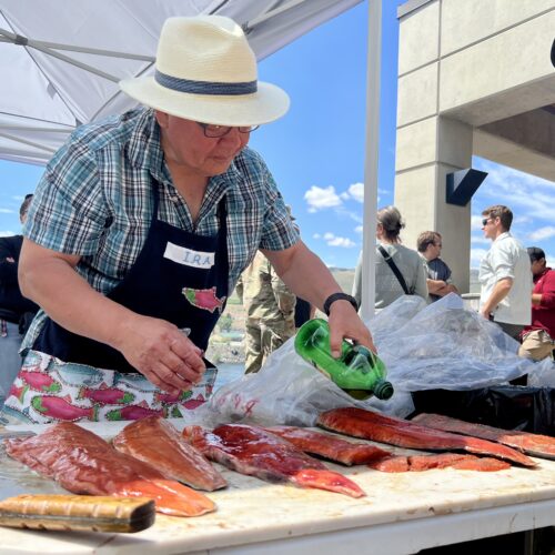 A man in a blue plaid shirt, black apron and white fedora holds a green bottle of olive oil above six pink filets of salmon. The filets are sitting on a white plastic table. A crowd of people stands behind him looking at a blue river.