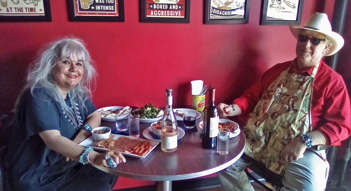 A woman with grey hair in a blue shirt and lots of turquoise jewlery sits at a table with a man in a red button down shirt, blue jeans, and a apron with horses and birds on it. They're sitting at a silver table. On the table are two wine bottles, a plate with cold cuts, a salad, some water glasses, and some other bowls. The wall behind them is bright red and has five cartoon paintings of cats drinking beer.