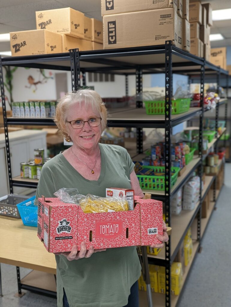 A woman with white hair and glasses holds a bright red box filled with bread, pasta, and canned food items. 