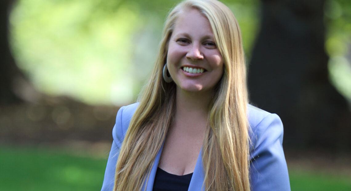 Portrait of Rebecca Scofield. She wears a periwinkle suit and black top with long blonde hair.