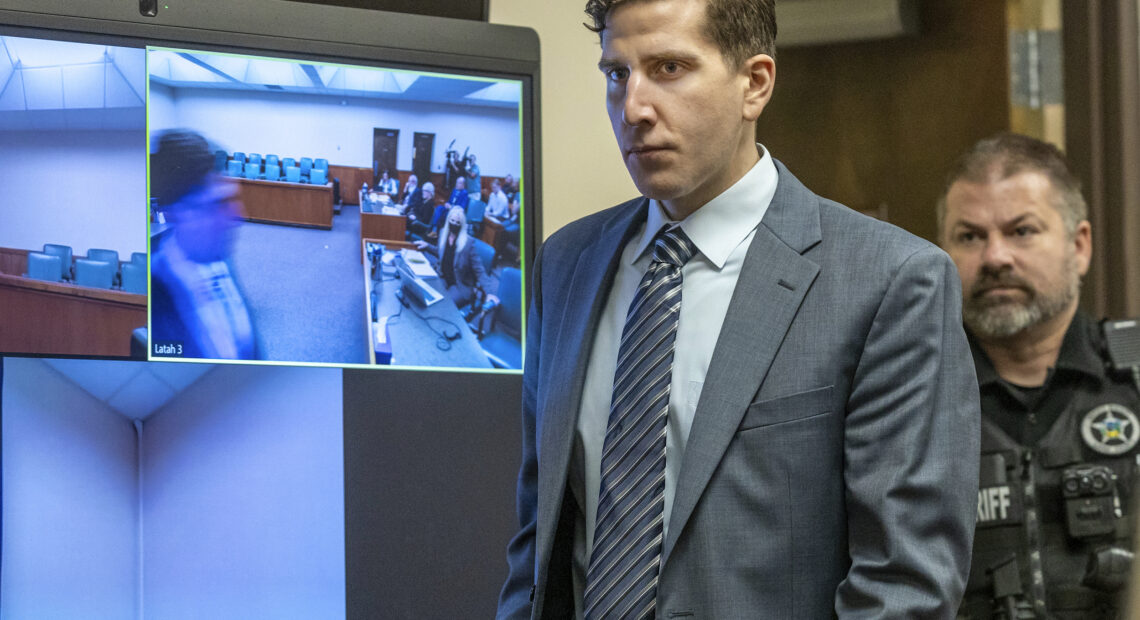 Bryan Kohberger wears a grey suit and striped grey tie as an officer in uniform walks behind him into the courtroom.