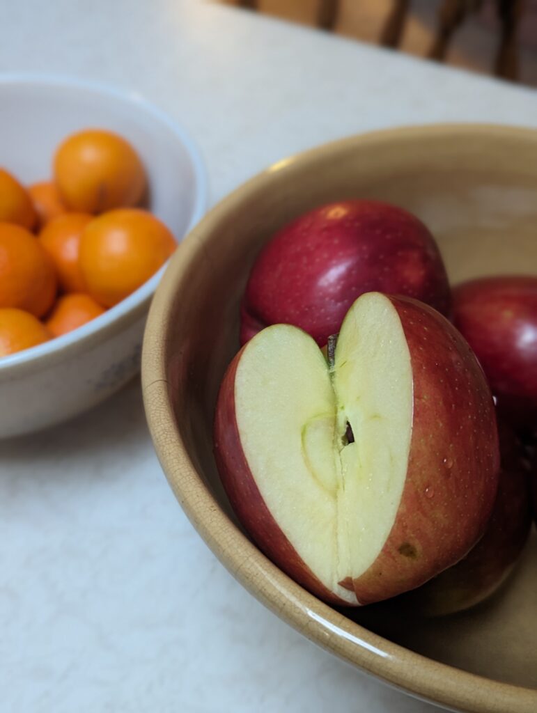 A sliced open Cosmic Crisp apple sits in a golden bowl next to a white bowl of small oranges.