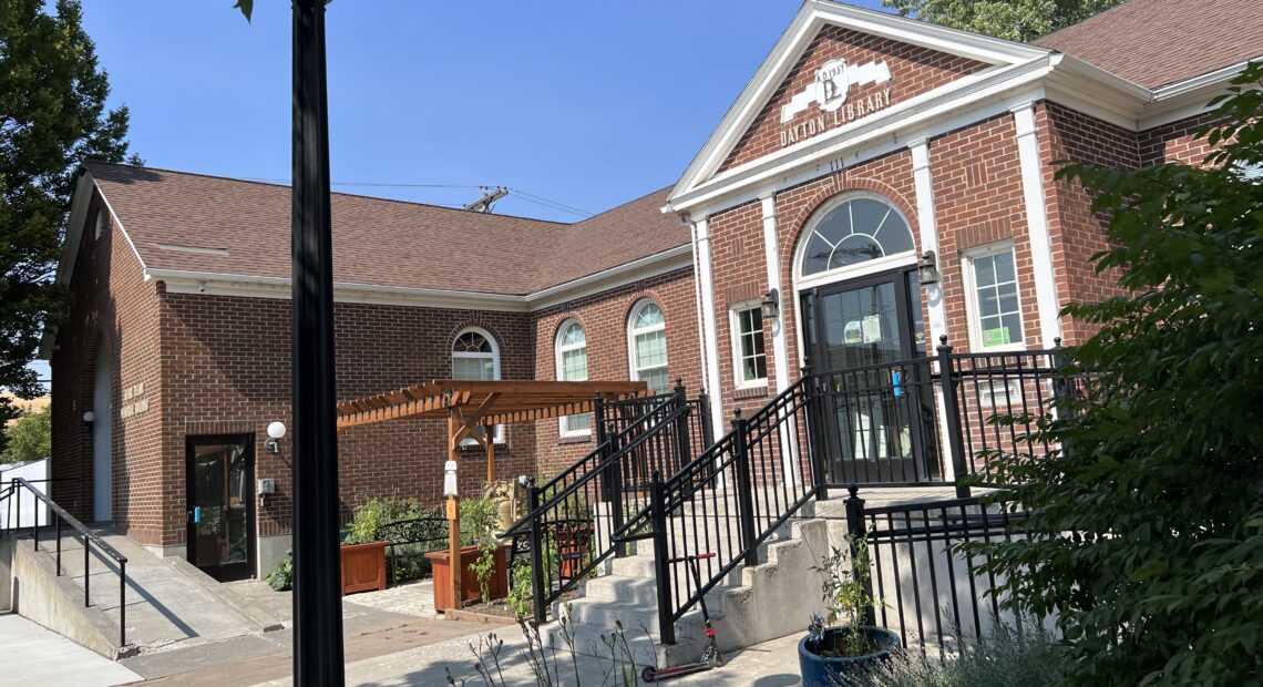 A red brick building with black stair railings. A small brown pergola is in front of the library. A few green plants are underneath the pergola. An evergreen tree is at the side of the red brick building. A black lamppost is in front of the building.