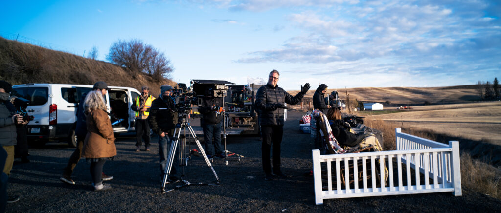 A camera is set up behind a white picket fence while crew members dressed in black fill the scene against a wheat field and blue sky.