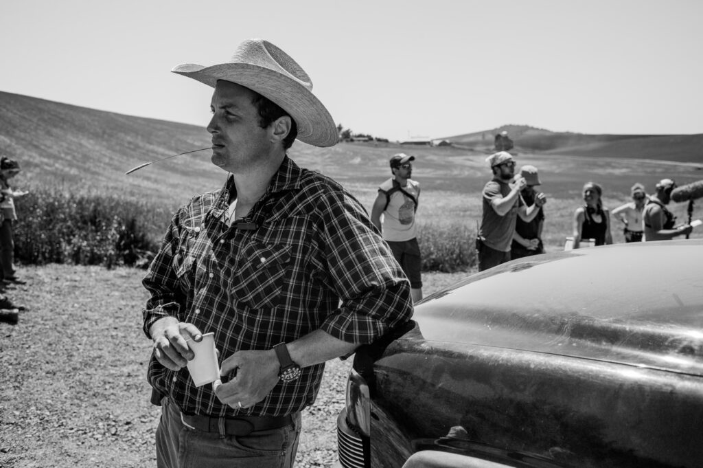 A man in a white cowboy hat and plaid shirt leans on a truck in front of a field in rural Washington. 