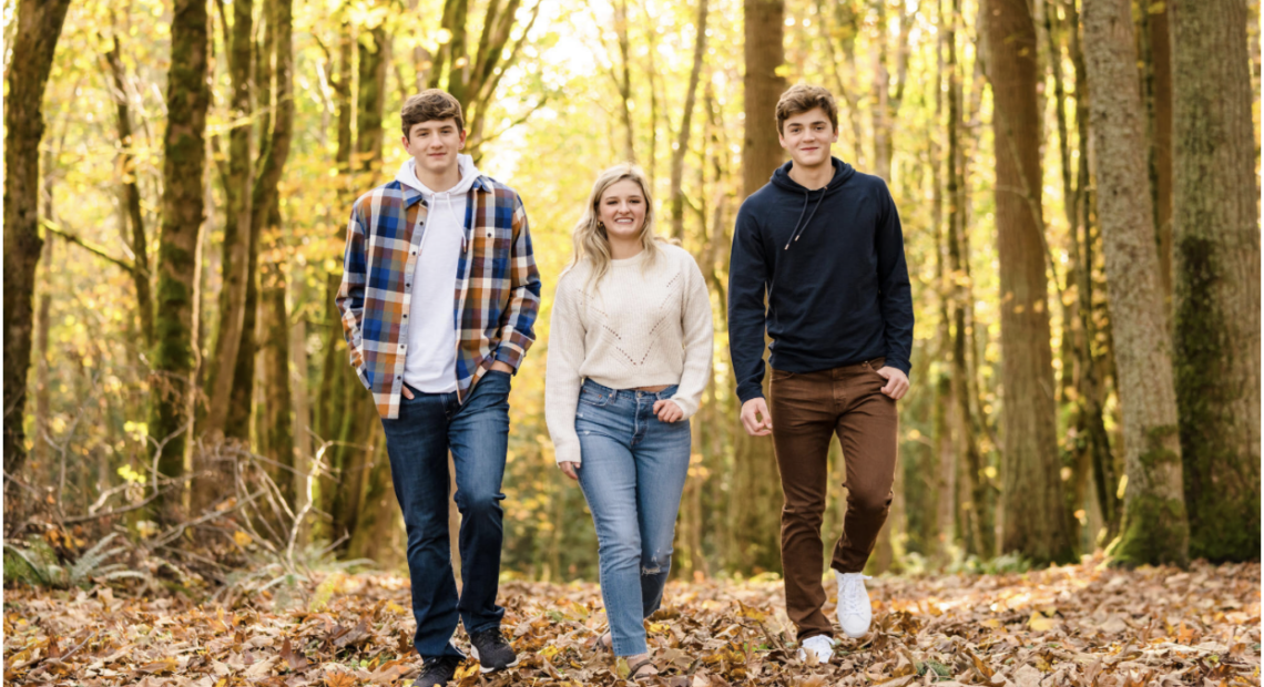 Two young men and a young woman in fall clothes stroll toward the camera between autumn trees.