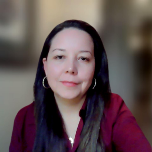 A portrait of Johanna Bejarano: she wears a long-sleeved red top, necklace and hooped earrings. She has long, dark, straight hair and is smiling toward the camera. The background is blurred.