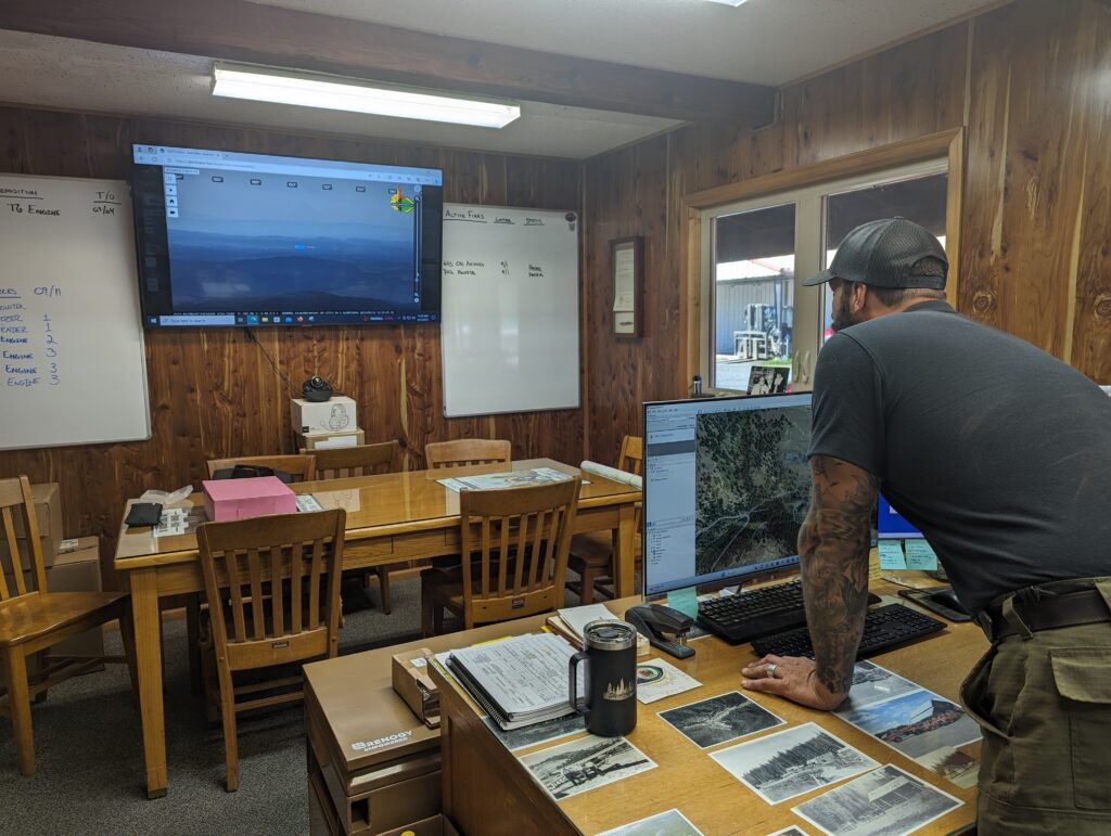 A man wit a sleeve tattoo and a black shirt moves the mouse on a computer screen showing a photo of the forest. 