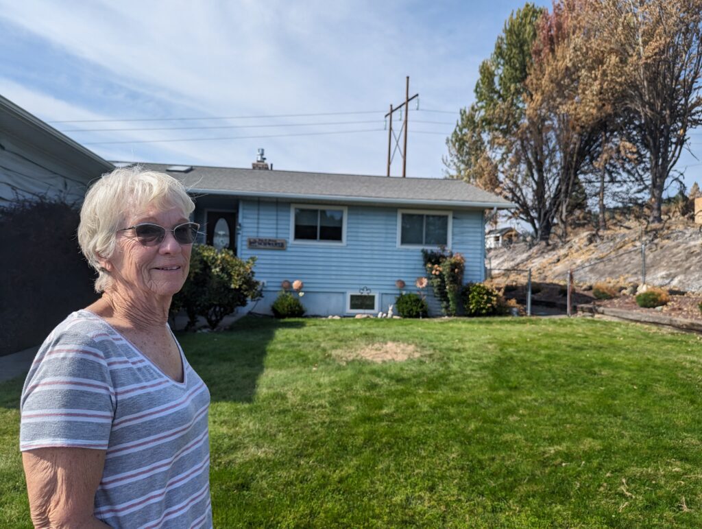 A woman with a white shirt and sunglasses stands in front of a blue house with a green lawn.