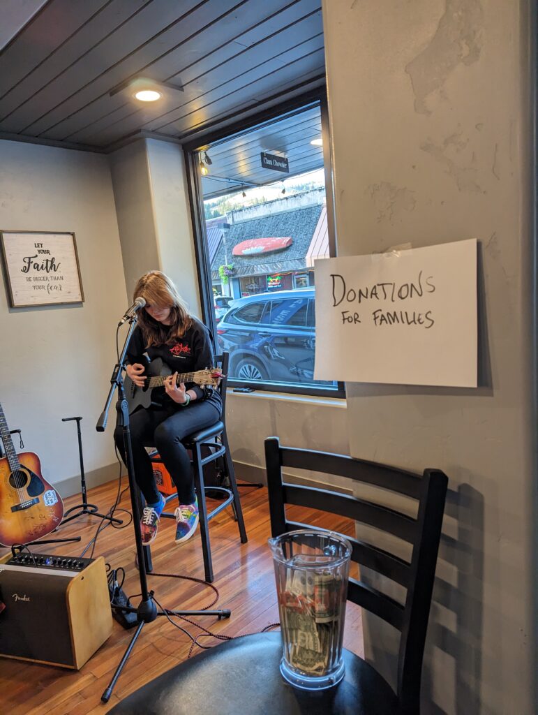 A girl with a guitar sits on a tall black chair in front of a microphone. A jar with "Donations for families" written above it sits nearby.