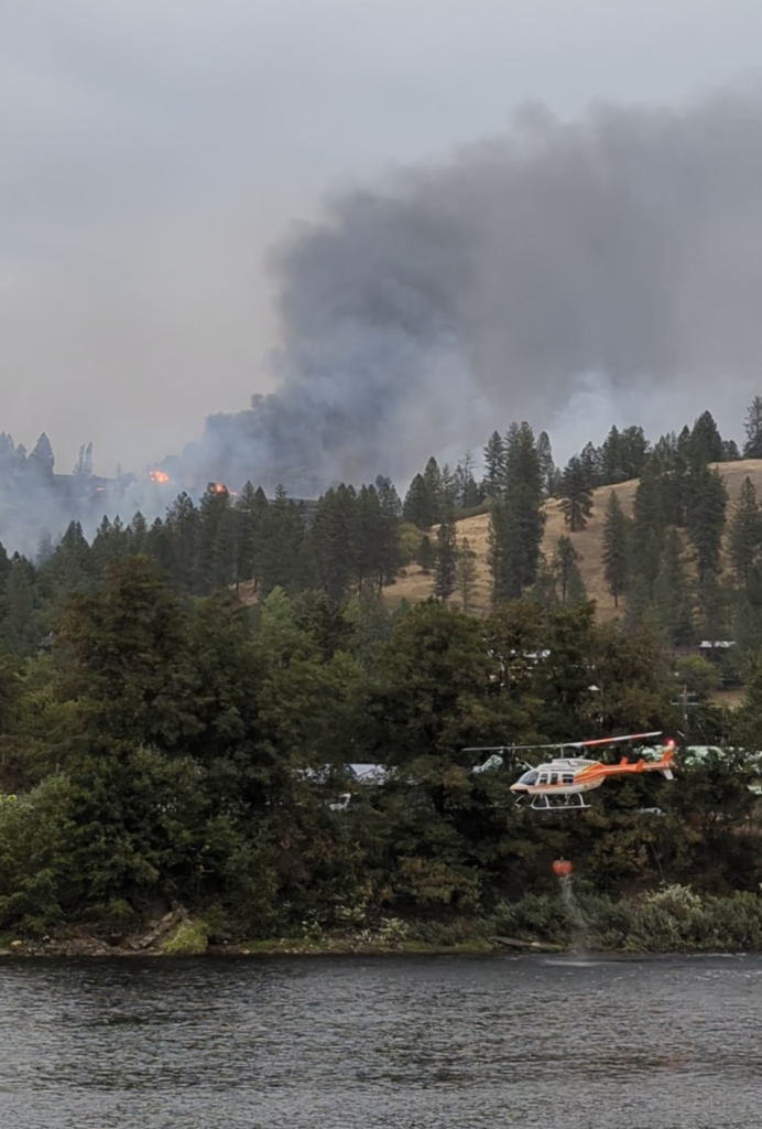 A helicopter scoops water with a bucket out of a river. A fire burns on a tree covered hill in the distance. 