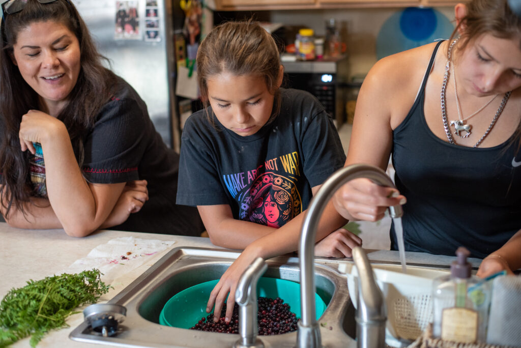 A mother stands on the left side in her kitchen, her arms resting on a counter top as her youngest daughter who is in her pre-teens looks down into a sink with a bowl of purple berries inside. The older, teenage daughter stands on the far right and is using the sink to run over a white strainer she holds in the water. She wears a silver necklace with a horse on it and a black tank top. The youngest daughter and mother wear black t-shirts. All three have long, dark hair and are focused on their tasks. The mother is smiling, her mouth open in conversation and black-framed glasses pushed up on her head.