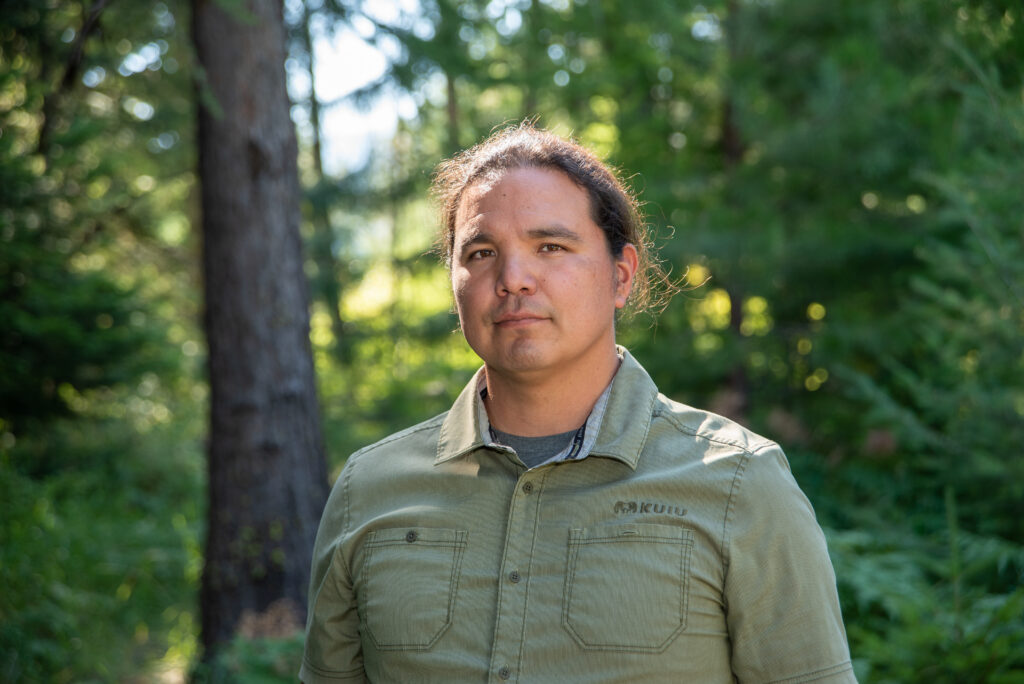 A man poses for a portrait facing the camera. He wears a green, button-down shirt and his hair is tied back. In the background, green pine trees are out of focus in a forest setting.