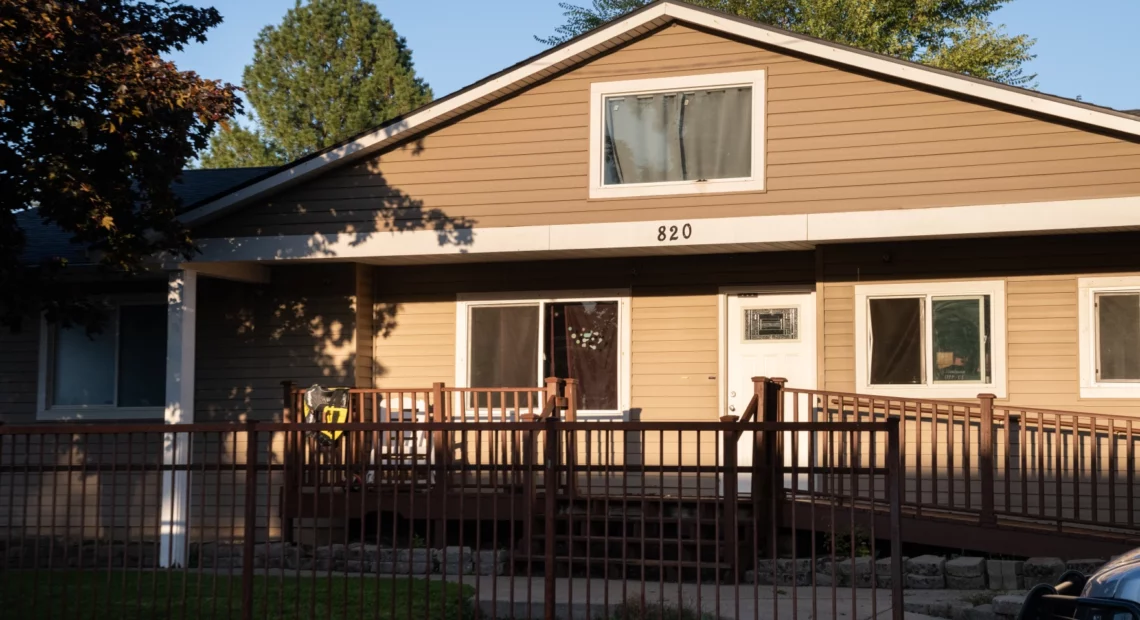 A brown, 2-story house is shown with blue skies in the background and green trees. Curtains in the house are closed and the numbers "820" are nailed into the center representing the address. A gate and brown fence with ramp and a few stairs go down to a small green grass yard in front.