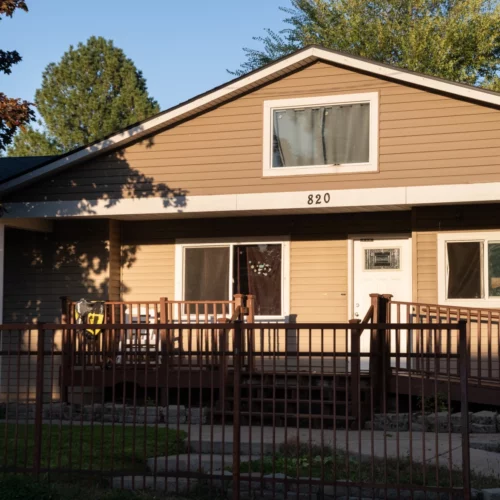 A brown, 2-story house is shown with blue skies in the background and green trees. Curtains in the house are closed and the numbers "820" are nailed into the center representing the address. A gate and brown fence with ramp and a few stairs go down to a small green grass yard in front.