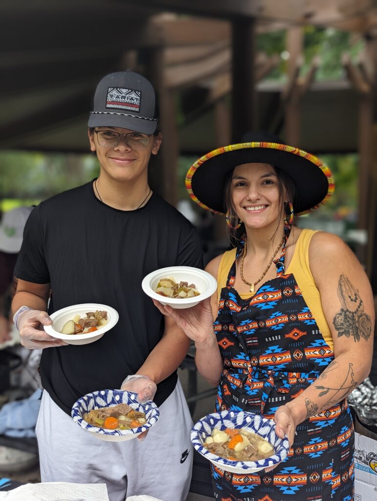 Two young tribal members in black hats hold bison and elk stew while smiling. 