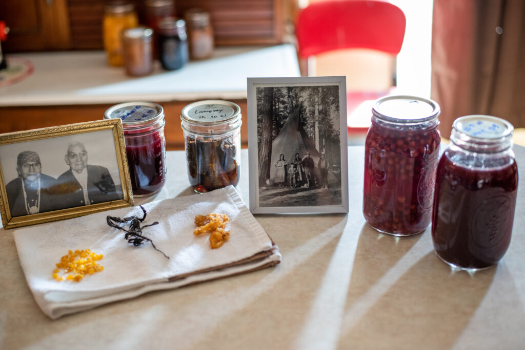 A golden framed photo of two elderly relatives of the Wolf family sits besides jars of canned First Foods and a display of dried, harvested foods. A silver-framed photo near the jars shows a historic scene of a young, Indigenous family standing outside a traditional house the family uses for hunting outings called a "tepee."