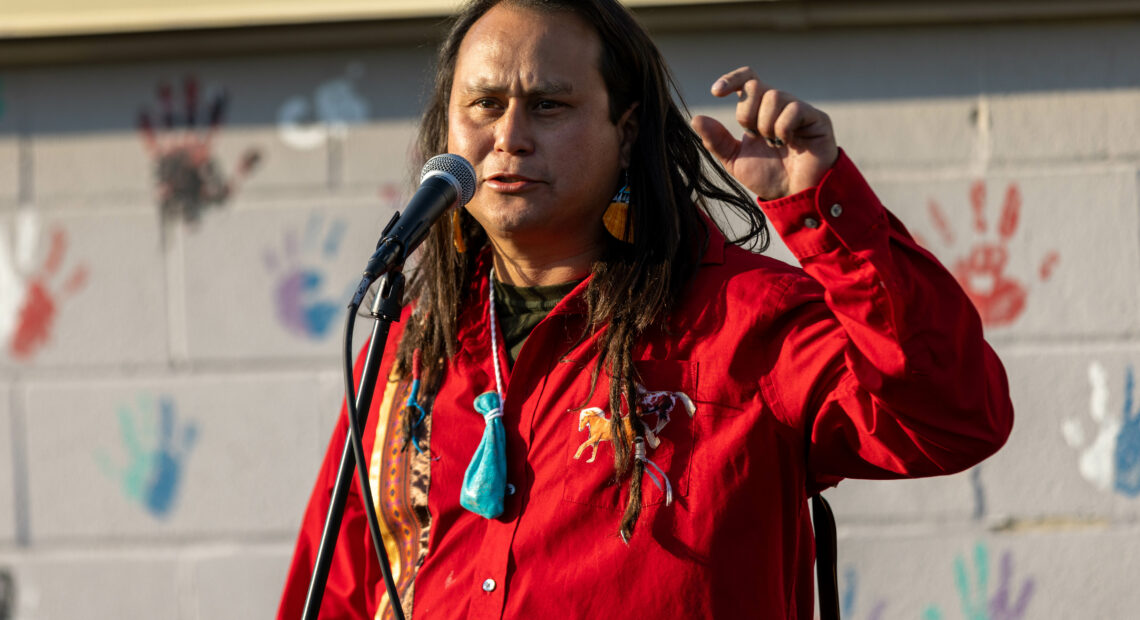 A Native American man with long dark hair in a red shirt stands in front of a brick wall with hand prints painted on it.