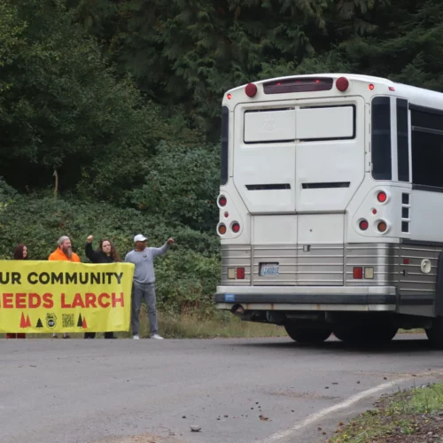 A handful of people stand holding a bright yellow banner that reads "Our Community Needs Larch" as the back of a bus is seen driving past them.