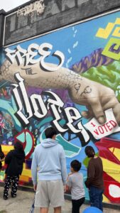 A brightly colored mural of a hand holding a sign that says "I voted" is covered with Free the Vote in white lettering. 